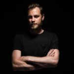 Portrait of a bearded man with an intense look standing in a dark room against a black background.