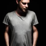 Portrait of a bearded man with an intense look standing in a dark room against a black background.